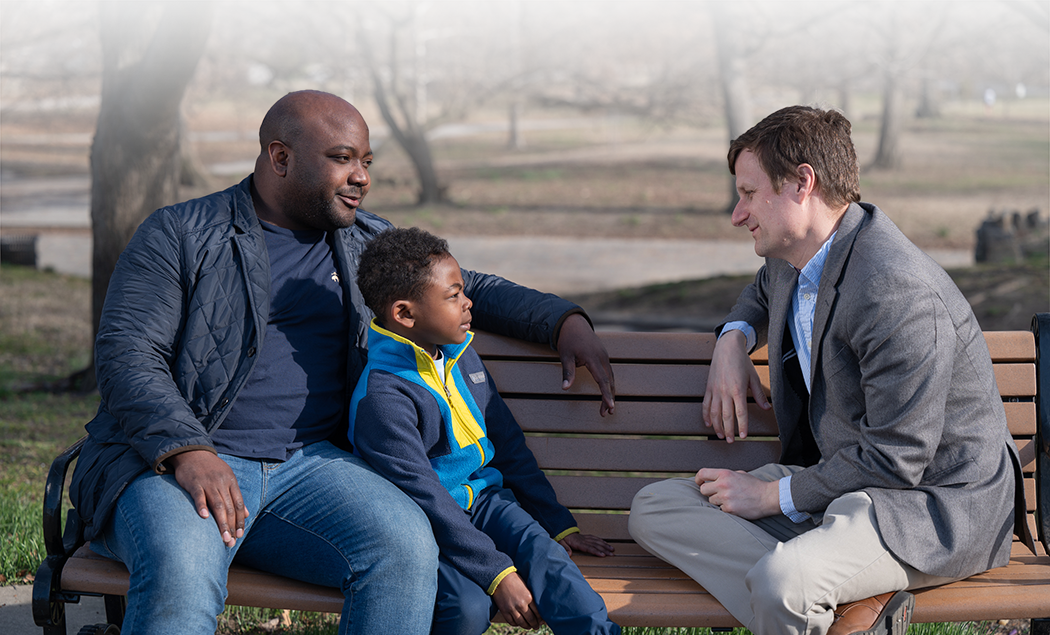 Mark Parker listens to a young Black boy and his father on a bench in Patterson Park